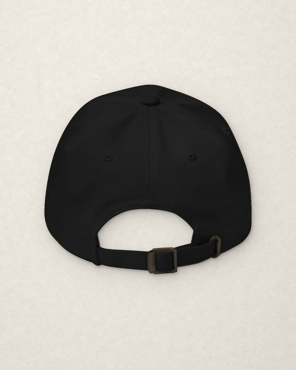 Back of the black dad cap on a warm off-white plaster surface, showing the plain rear panels and the adjustable fabric strap with a metal slide buckle. No embroidery on this side.