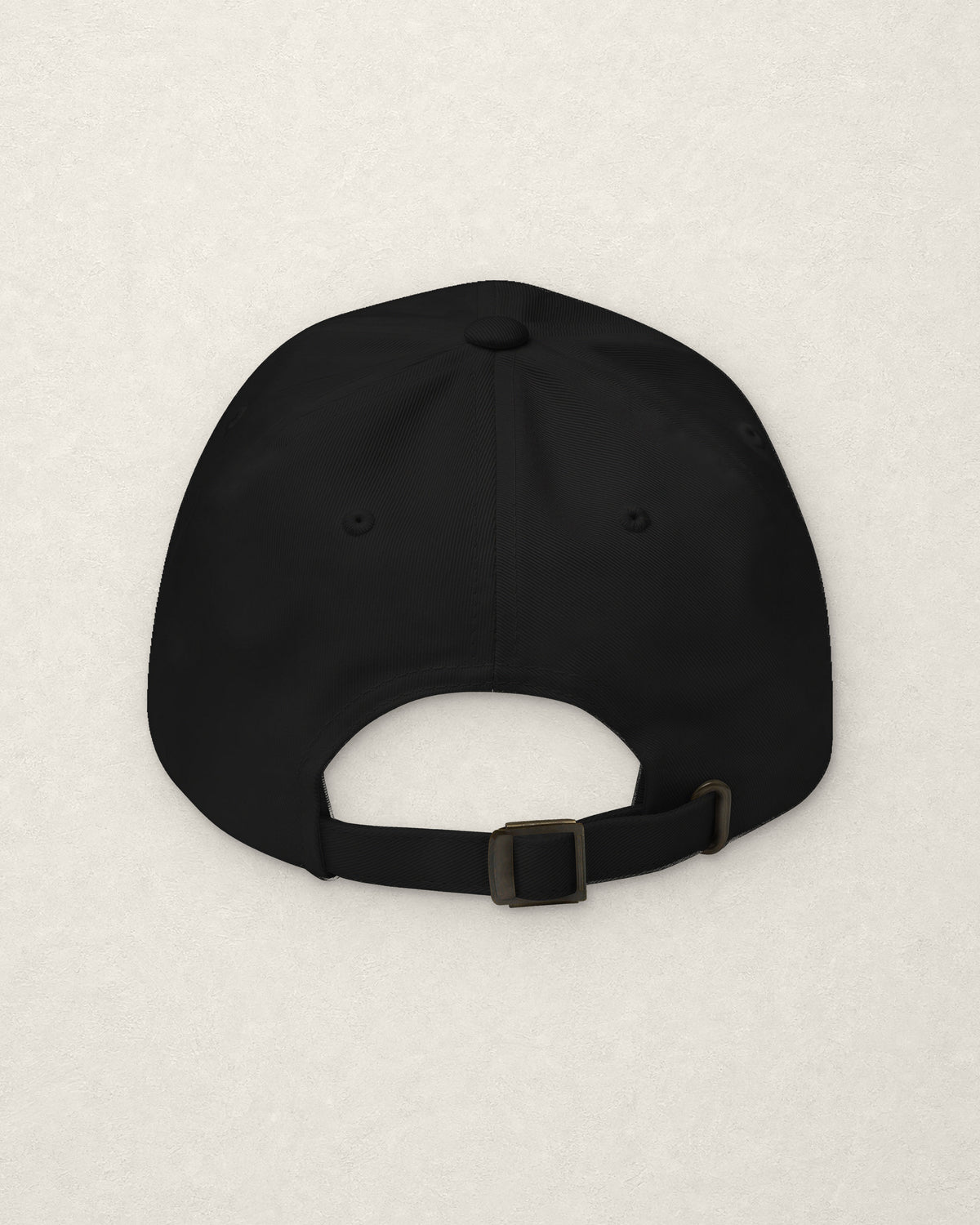 Back of the black dad cap on a warm off-white plaster surface, showing the plain rear panels and the adjustable fabric strap with a metal slide buckle. No embroidery on this side.