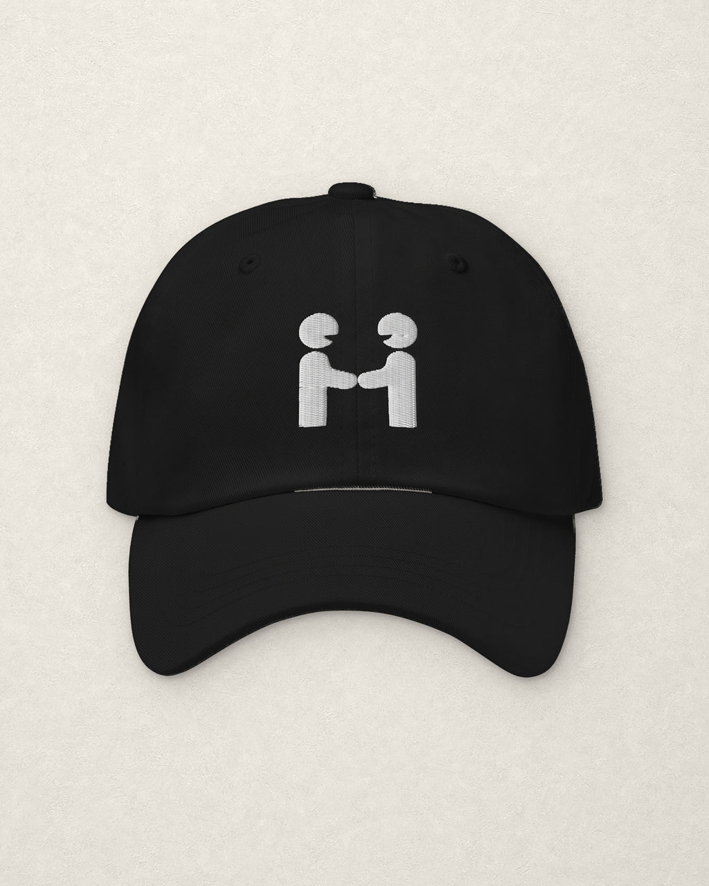 Straight-on studio view of the black unstructured dad cap on a warm off-white plaster surface, with the Human-H embroidered on the front panel in off-white thread: two figures shaking hands.