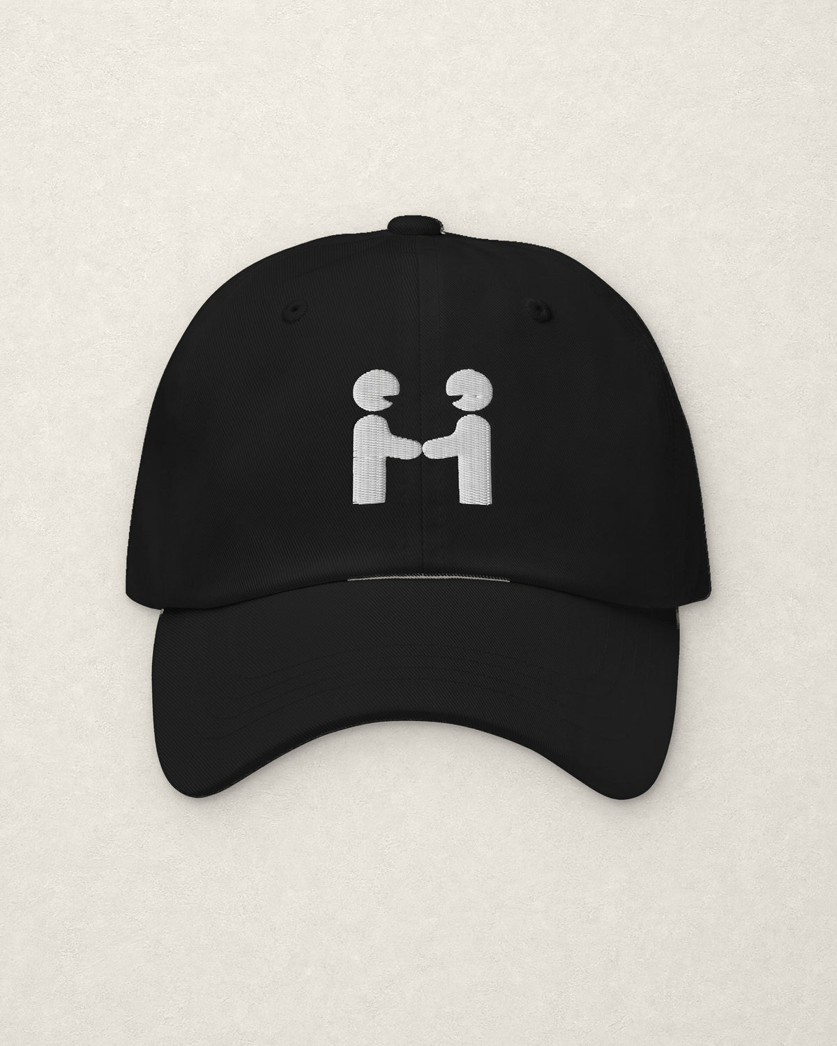 Straight-on studio view of the black unstructured dad cap on a warm off-white plaster surface, with the Human-H embroidered on the front panel in off-white thread: two figures shaking hands.