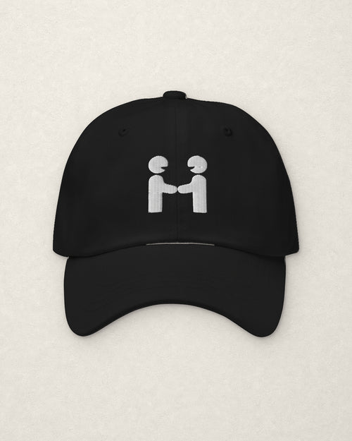 Straight-on studio view of the black unstructured dad cap on a warm off-white plaster surface, with the Human-H embroidered on the front panel in off-white thread: two figures shaking hands.