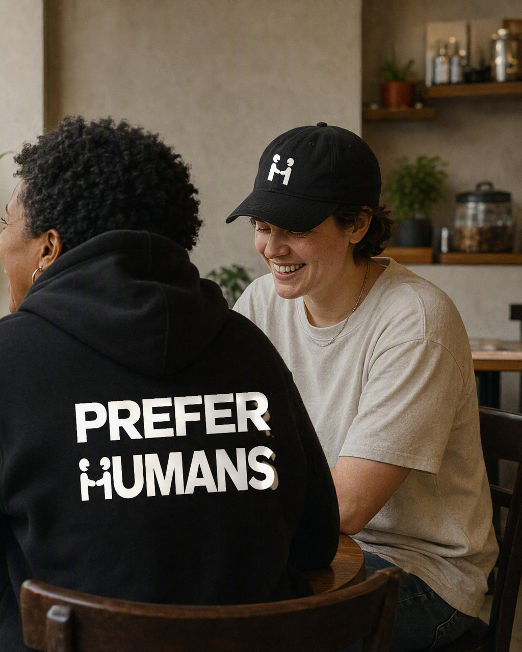 Human-H cap worn at a cafe window table, beside a friend in the PREFER HUMANS hoodie with the back print visible.