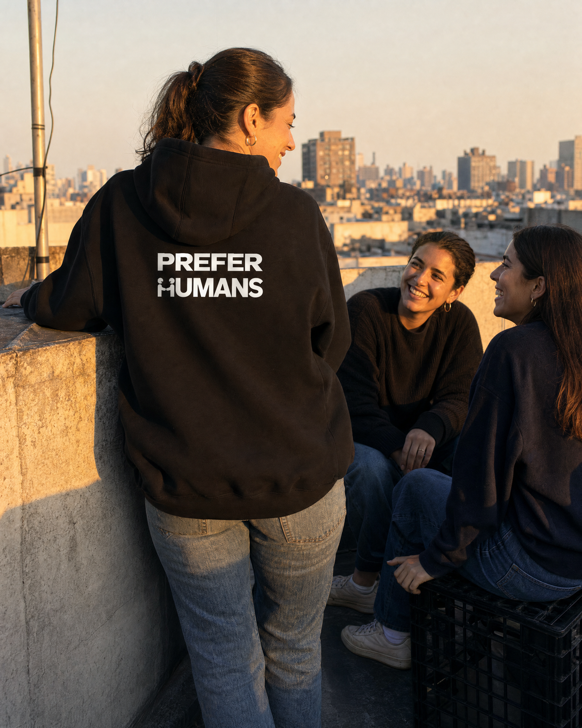Two people on a rooftop at golden hour, one wearing the PREFER HUMANS hoodie seen from behind