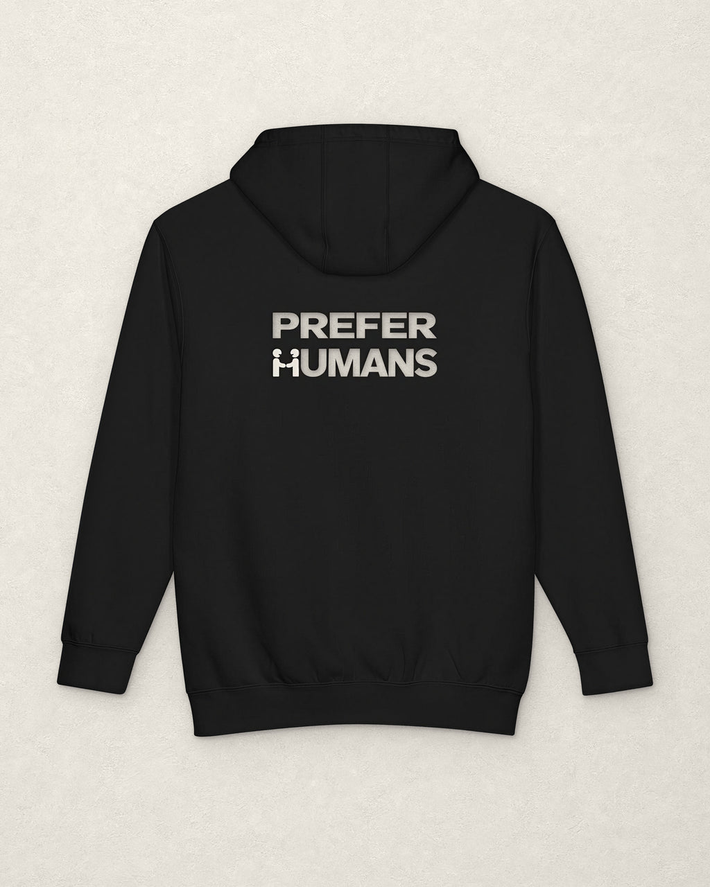Back of the black PREFER HUMANS hoodie on a warm off-white plaster surface, printed below the hood in off-white with the words PREFER HUMANS on two lines, the H of HUMANS formed by two figures shaking hands.