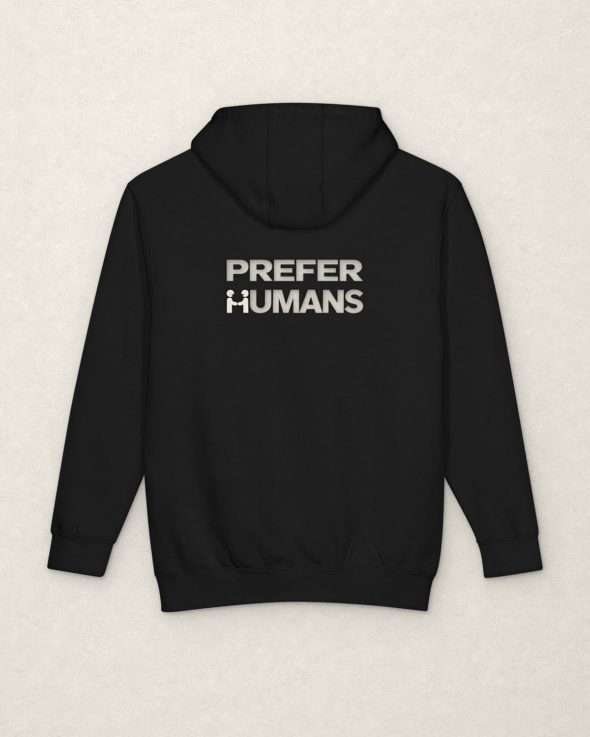 Back of the black PREFER HUMANS hoodie on a warm off-white plaster surface, printed below the hood in off-white with the words PREFER HUMANS on two lines, the H of HUMANS formed by two figures shaking hands.