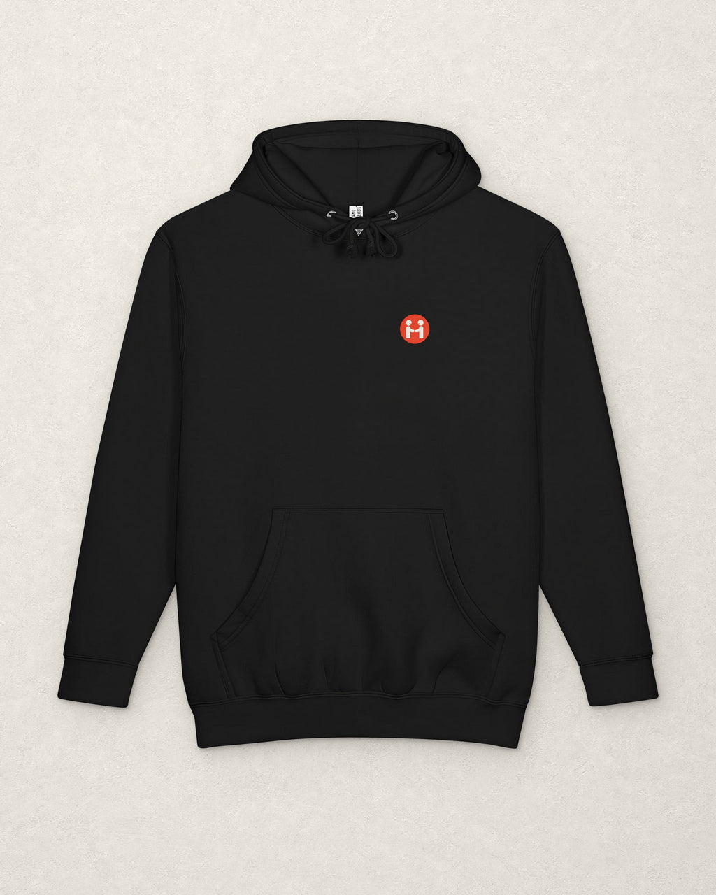 Front of the black PREFER HUMANS hoodie laid flat on a warm off-white plaster surface, with drawcords and a kangaroo pocket, and a small round red Human Signal badge showing two figures shaking hands on the left chest.