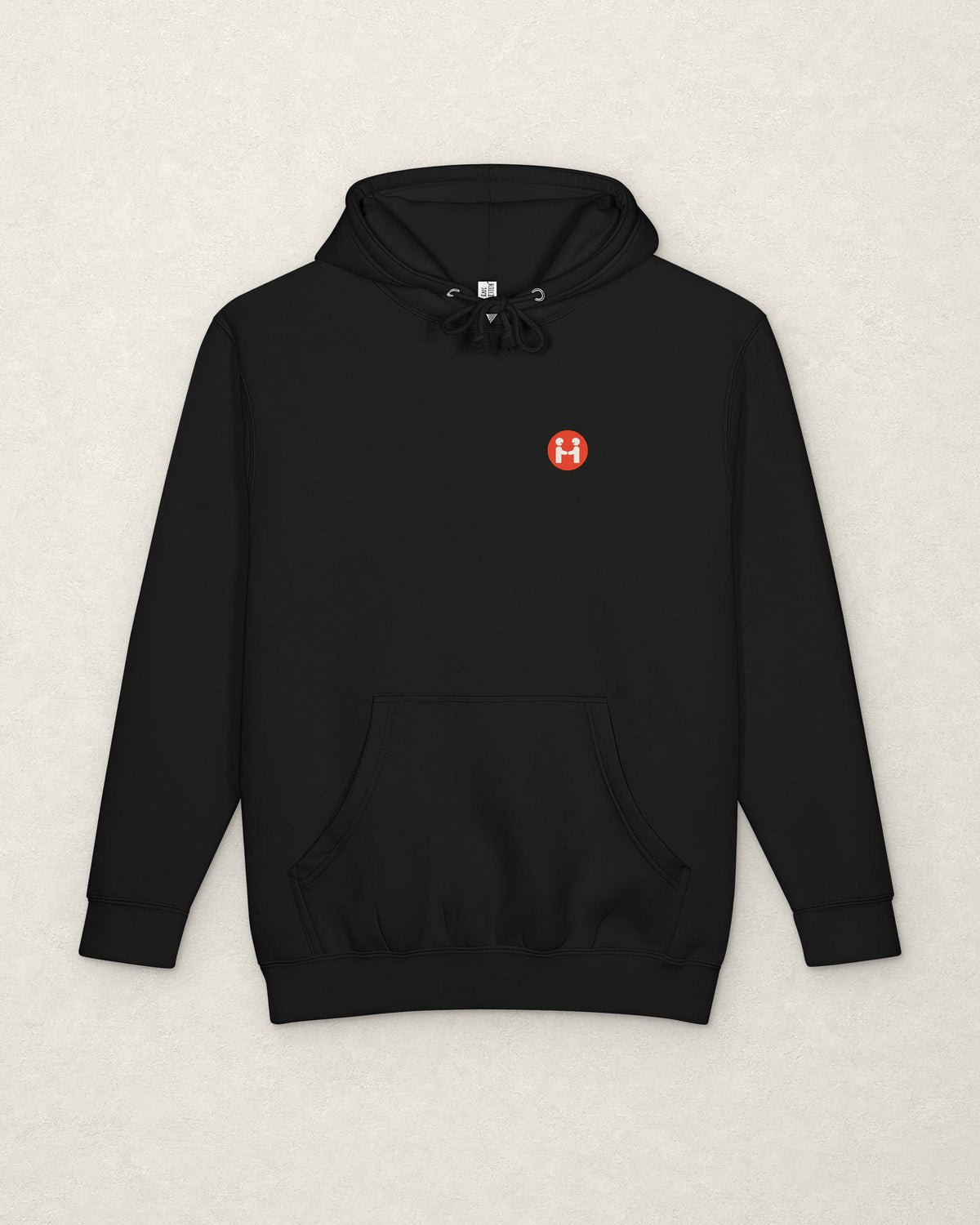 Front of the black PREFER HUMANS hoodie laid flat on a warm off-white plaster surface, with drawcords and a kangaroo pocket, and a small round red Human Signal badge showing two figures shaking hands on the left chest.