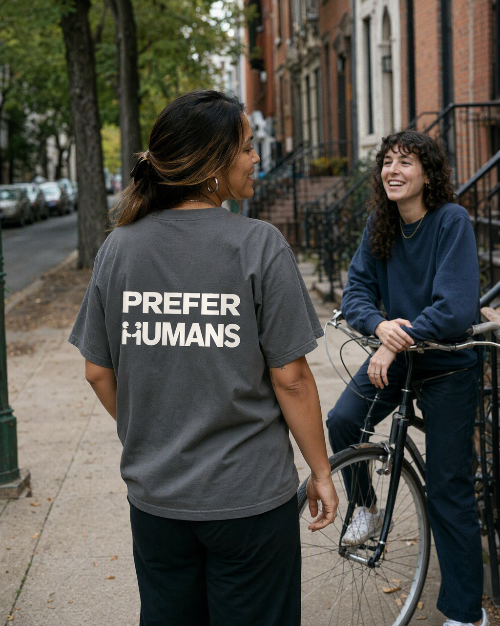 PREFER HUMANS tee back print on a leafy residential sidewalk, talking with a friend on a bike
