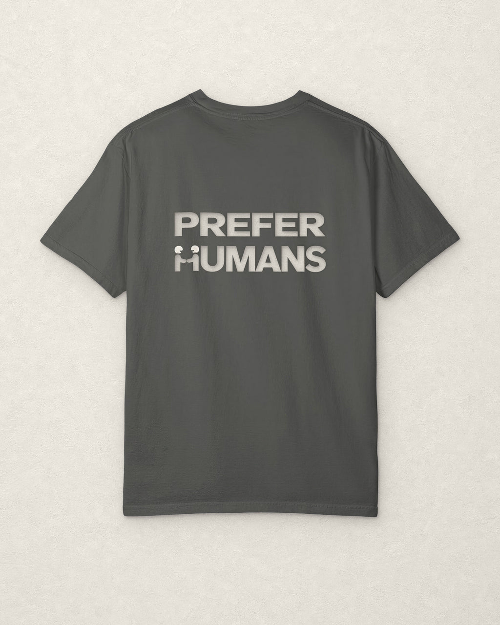 Back of the charcoal PREFER HUMANS tee on a warm off-white plaster surface, printed across the shoulders in off-white with the words PREFER HUMANS on two lines, the H of HUMANS formed by two figures shaking hands.