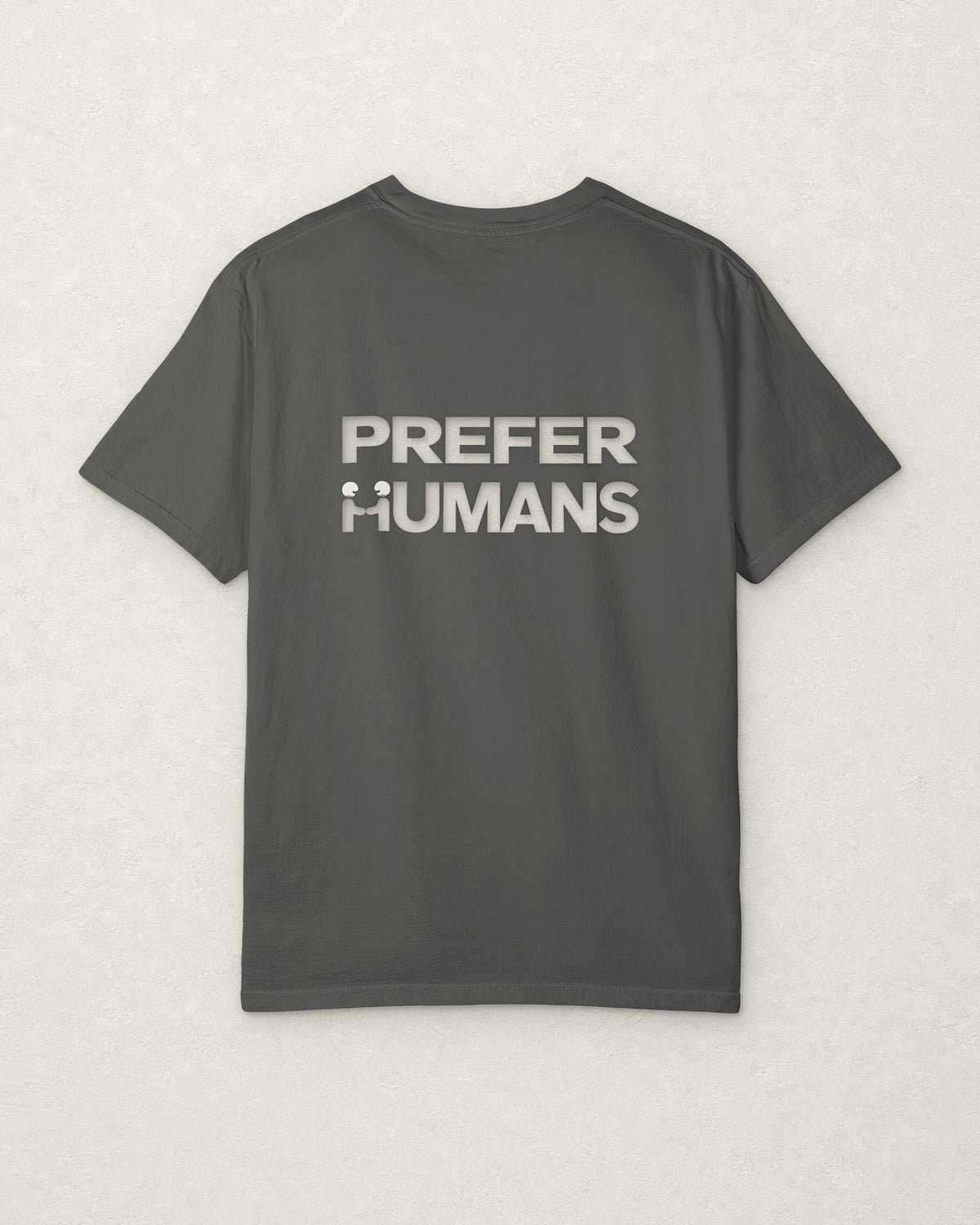 Back of the charcoal PREFER HUMANS tee on a warm off-white plaster surface, printed across the shoulders in off-white with the words PREFER HUMANS on two lines, the H of HUMANS formed by two figures shaking hands.