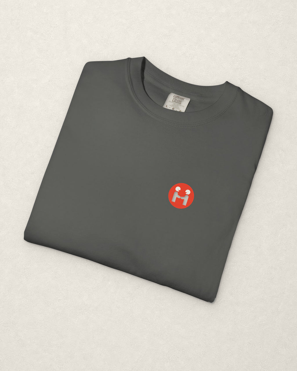 The charcoal PREFER HUMANS tee folded on a warm off-white plaster surface, with the red Human Signal badge and the Comfort Colors label visible.