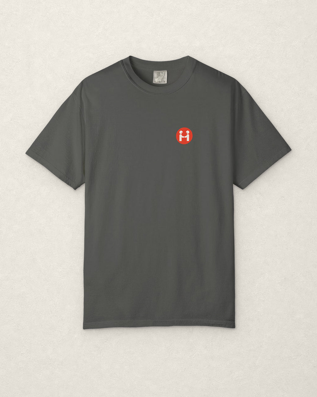 Front of the charcoal PREFER HUMANS tee laid flat on a warm off-white plaster surface, showing the small round red Human Signal badge with two off-white figures shaking hands on the left chest, and the Comfort Colors neck label.