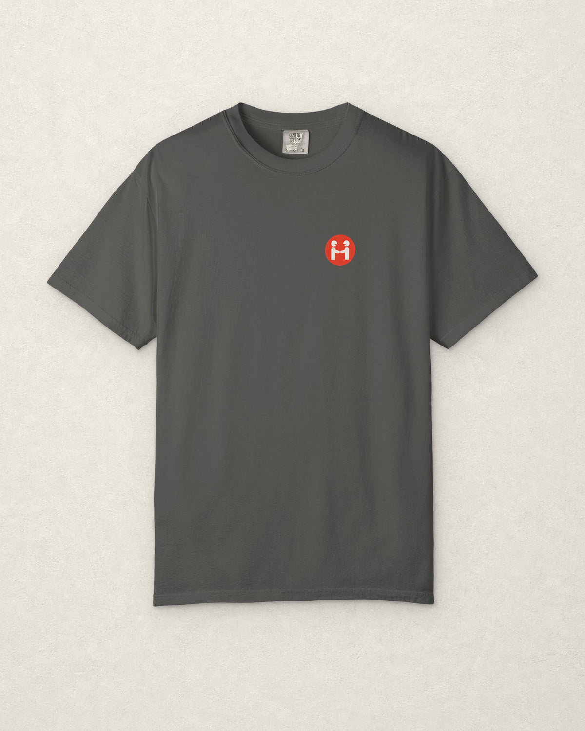 Front of the charcoal PREFER HUMANS tee laid flat on a warm off-white plaster surface, showing the small round red Human Signal badge with two off-white figures shaking hands on the left chest, and the Comfort Colors neck label.